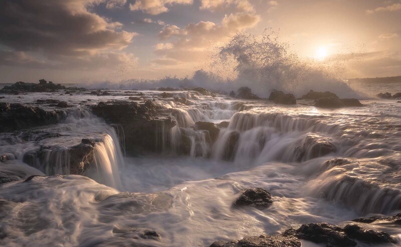 Powerful ocean waves crash against rocky coastline at sunrise, creating dramatic spray and turbulent white water.