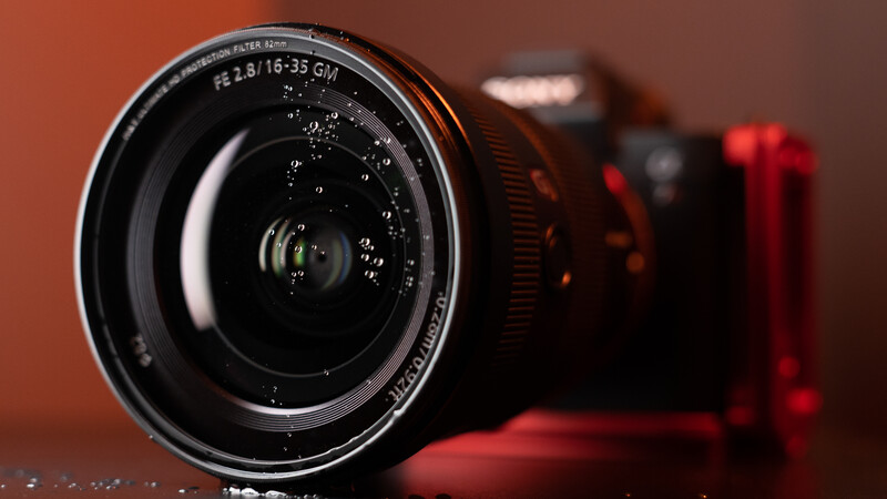 Wide-angle camera lens in sharp focus against warm orange background with blurred camera body behind.