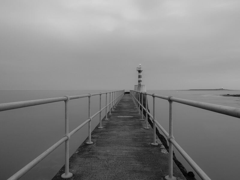 Long wooden pier with metal railings extending toward a distant lighthouse under overcast sky.