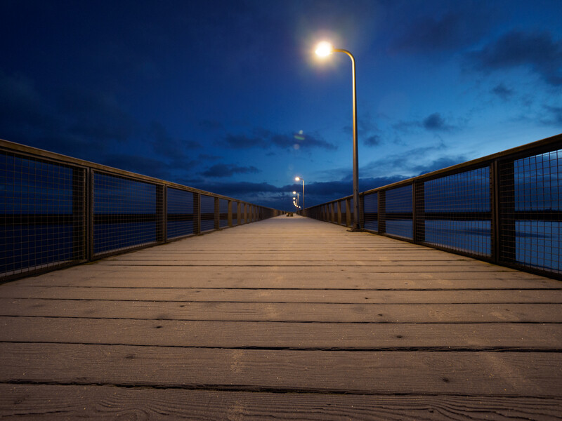 Wooden pier extending into calm water at twilight, lit by a tall street lamp against a deep blue sky.
