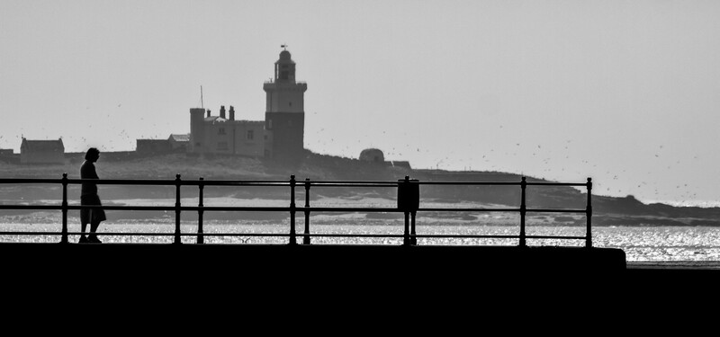 Silhouetted figures on a waterfront promenade with a lighthouse and coastal buildings across the water.