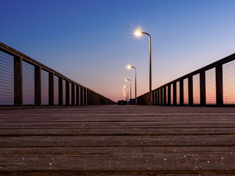 Empty wooden boardwalk stretching toward the horizon at dusk, lined with metal railings and street lamps.
