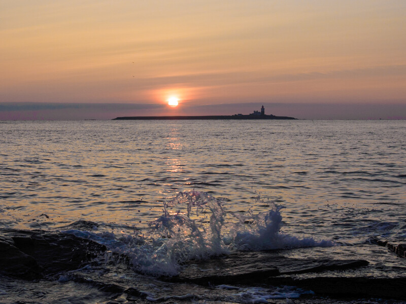 Ocean waves breaking on a beach at sunset, with a distant lighthouse silhouetted against the golden sun on the horizon.