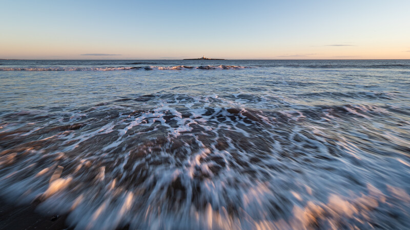 Ocean waves with motion blur rolling onto a beach at dusk, with distant headlands visible on the horizon.