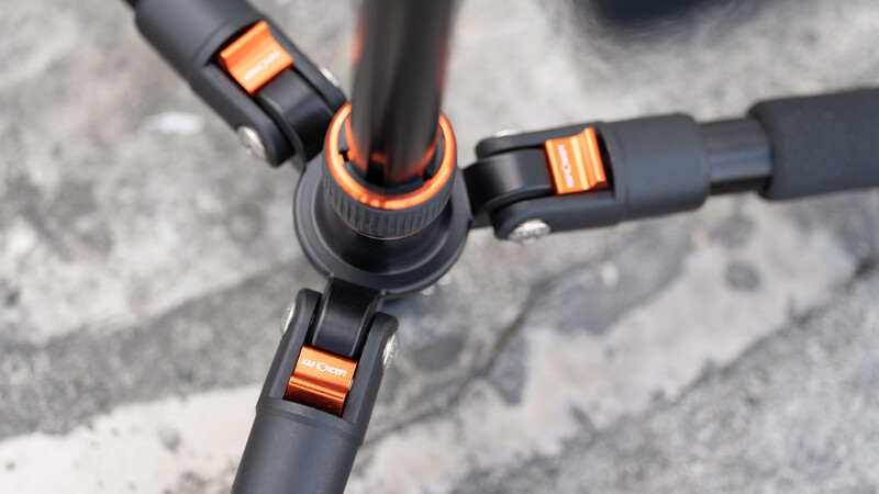 Close-up of a tripod head with three black legs and orange accent bands against concrete.