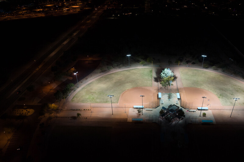 Aerial nighttime view of an illuminated baseball field with clay infield and grass outfield.