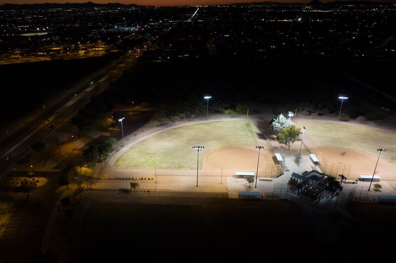 Nighttime aerial view of an illuminated sports field surrounded by dark landscape and distant city lights.