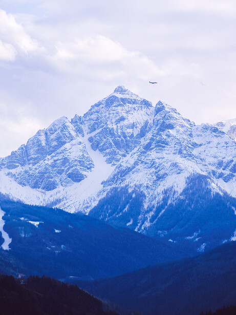 Snow-covered mountain peak with forested slopes under partly cloudy sky.