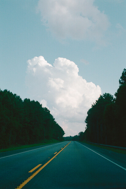 Straight highway with yellow center line stretching toward cumulus clouds on blue sky, tree-lined road.
