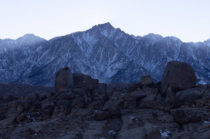 Rocky desert boulders in foreground with snow-covered mountain range at twilight.