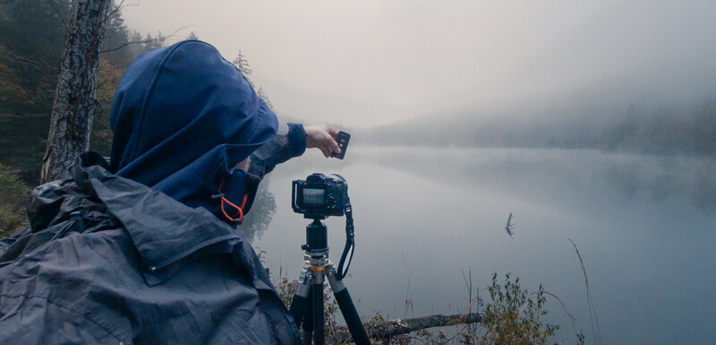 Photographer in blue jacket operating camera on tripod by misty lakeside at dawn.