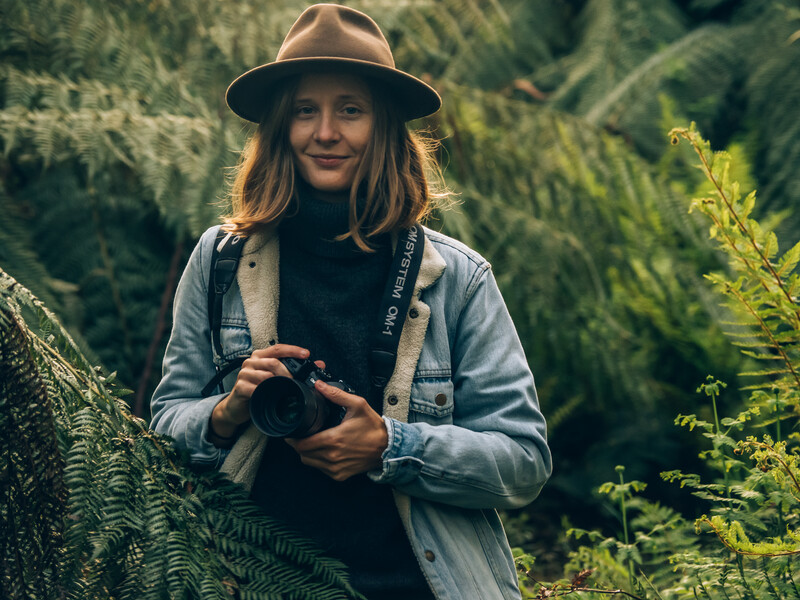 Woman in denim shirt and fedora holding camera in forested setting.