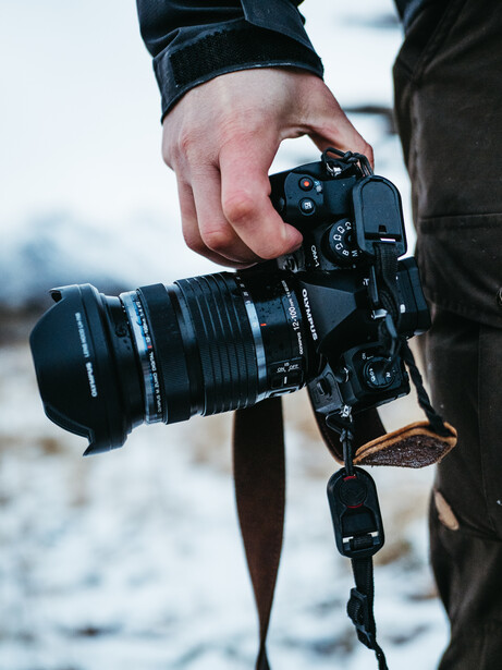 Photographer holding a mirrorless camera with telephoto lens attached, wearing a camera strap outdoors.