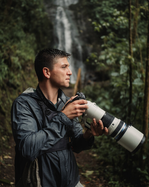 Male photographer holding a telephoto lens while looking toward a distant waterfall in a lush forest.