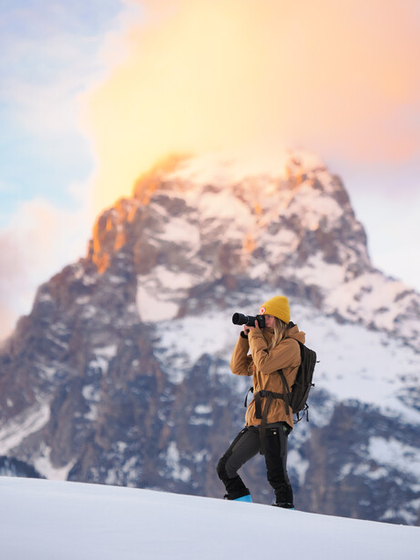 Backpacker in yellow jacket hiking across snow toward a snow-covered mountain peak at sunrise.