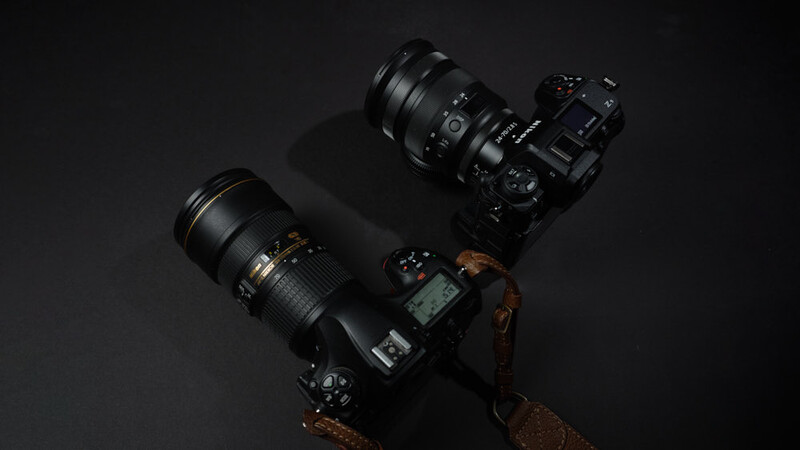 Two DSLR cameras with telephoto lenses arranged on a dark surface with dramatic side lighting.