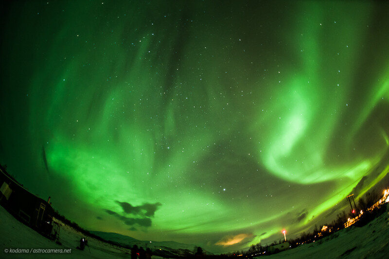 Vibrant green aurora borealis dancing across night sky with stars and illuminated landscape below.