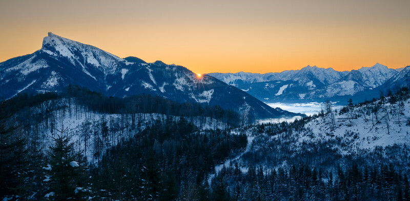 Snow-covered mountain range with forested slopes and a frozen lake visible in the valley below at twilight.