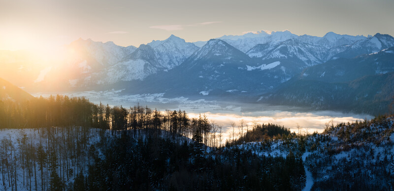 Snow-covered forested mountains with golden sunrise light breaking through mist in a valley below.
