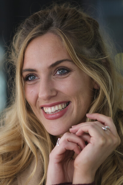 Woman with long blonde hair smiling at camera with hands near face.