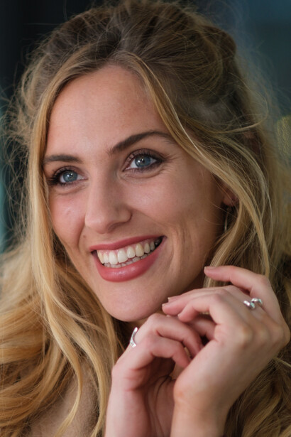 Woman with blonde hair smiling at camera with hands near face.