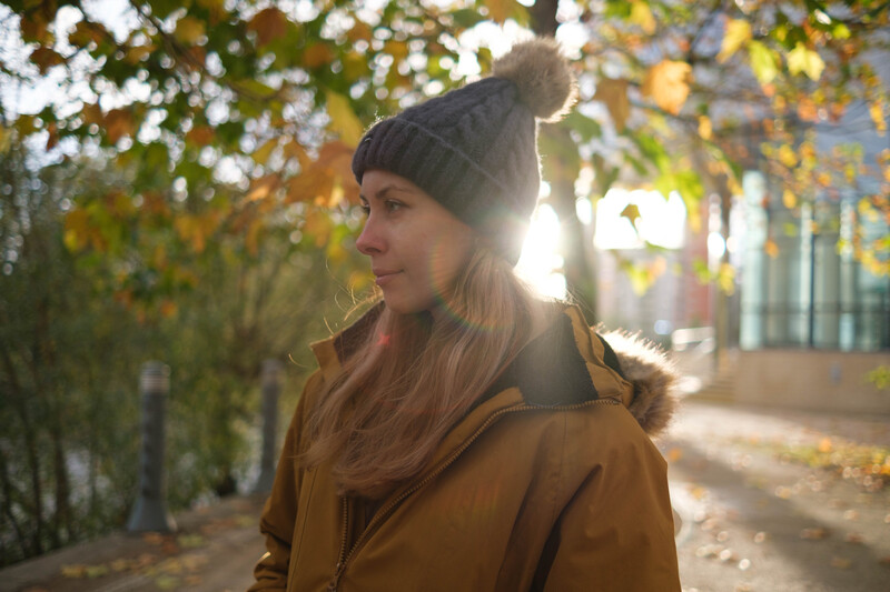 Woman in mustard jacket and knit beanie looking over shoulder in dappled autumn light beneath trees.