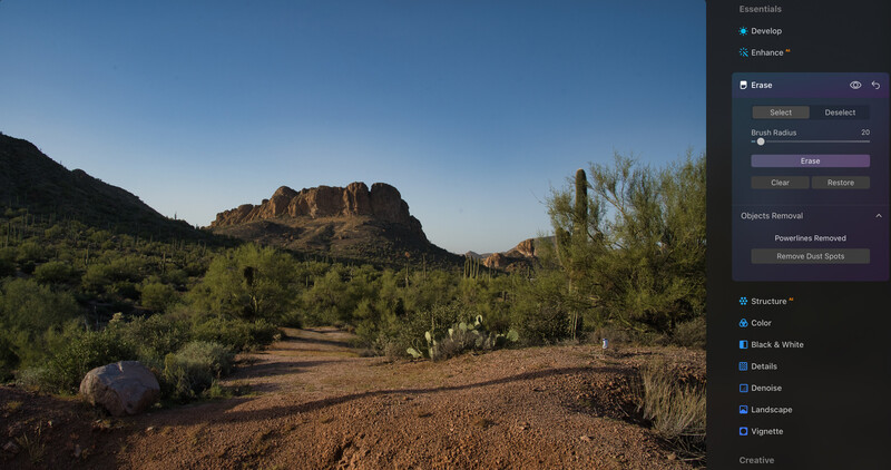 Desert landscape with rocky mountain formation surrounded by saguaro cacti and desert vegetation under clear blue sky.