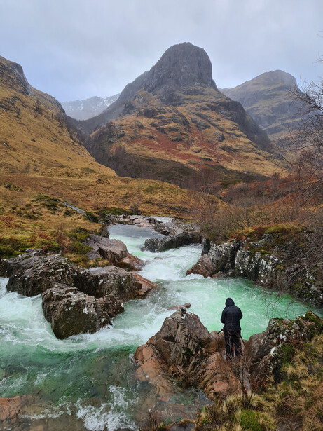Hiker standing on rocky outcrop overlooking turquoise glacial river and mountains.