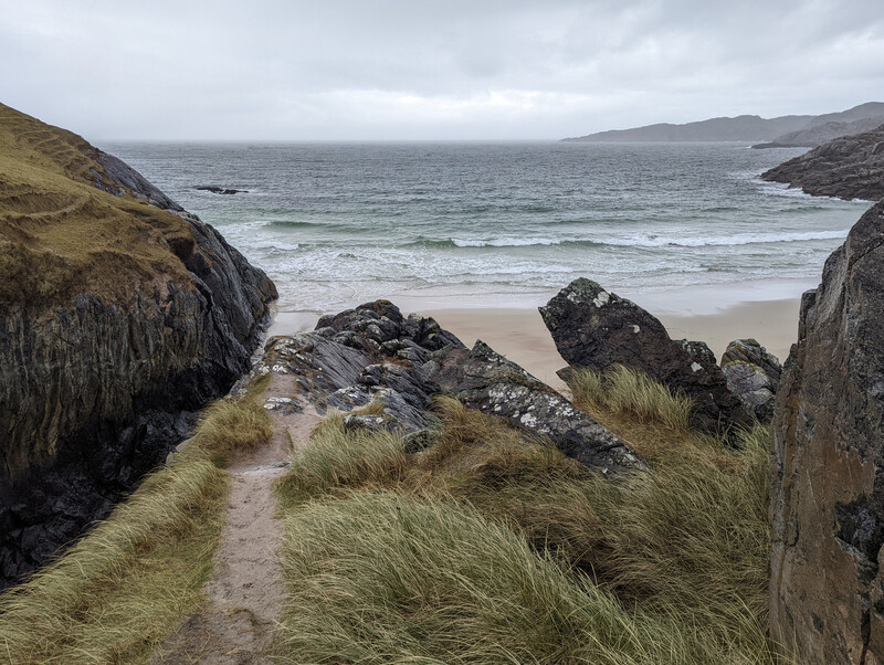 Coastal path with tufts of grass leading toward a beach framed by dark rocky outcrops.