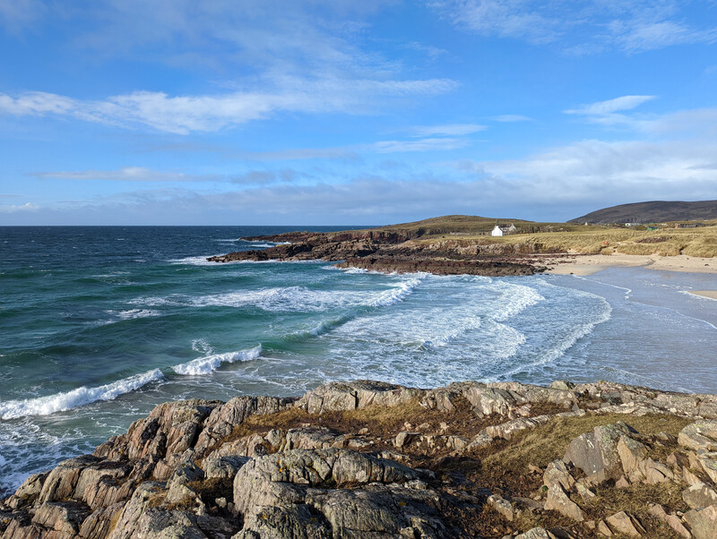 Coastal landscape with rocky foreground, sandy beach, breaking waves, and grassy headlands under blue sky.