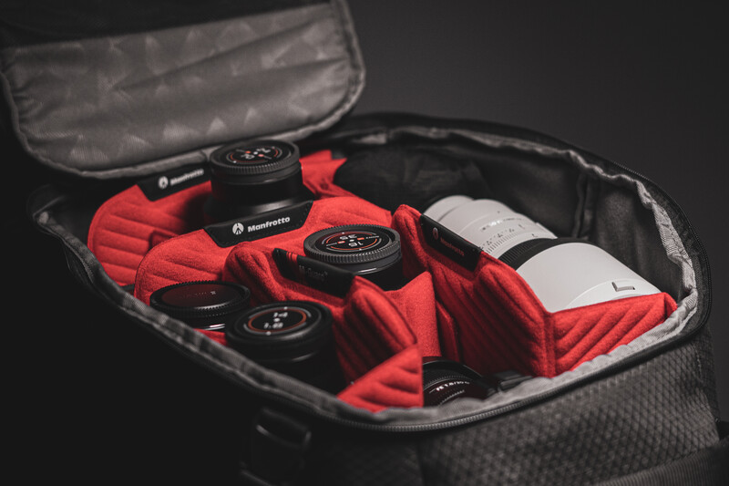 Open camera gear bag displaying red padded dividers, black lens caps, and white lens tucked inside.
