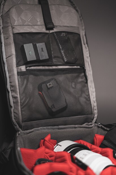 Open camera backpack displaying organized gear compartments with camera body, lens, accessories, and red fabric visible at bottom.