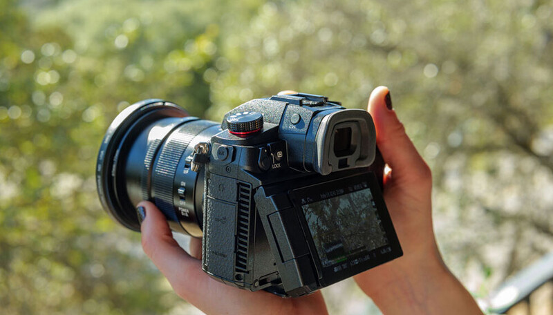 Medium format camera with articulating LCD screen held in hands outdoors.
