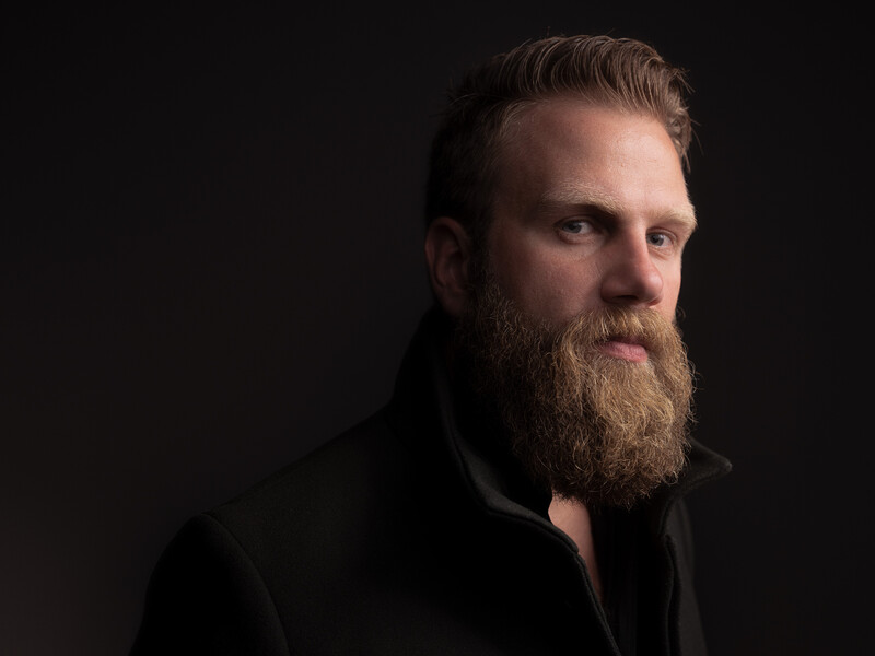 Bearded man with styled hair wearing black shirt against dark background.