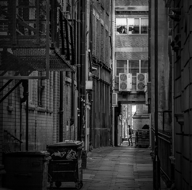Narrow urban alley between brick buildings with industrial windows and a single figure walking in the distance.