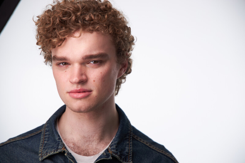 Young man with curly auburn hair wearing a dark denim shirt against a neutral white background.