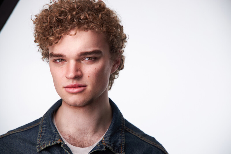 Young man with curly auburn hair wearing a dark denim shirt against a neutral white background.
