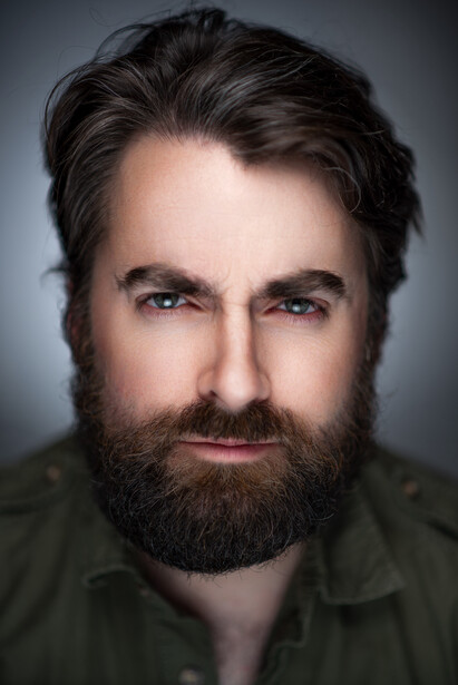 Man with dark hair and full beard wearing olive green shirt against neutral gray background.