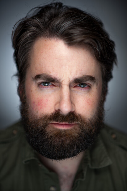 Man with dark hair and full beard wearing olive green shirt against neutral gray background.