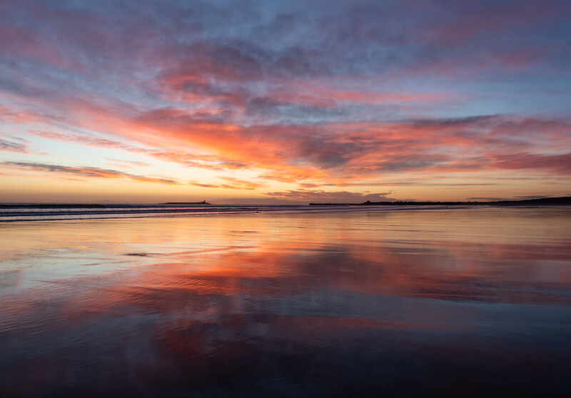 Vibrant sunset reflected in still wet sand on a beach at low tide.