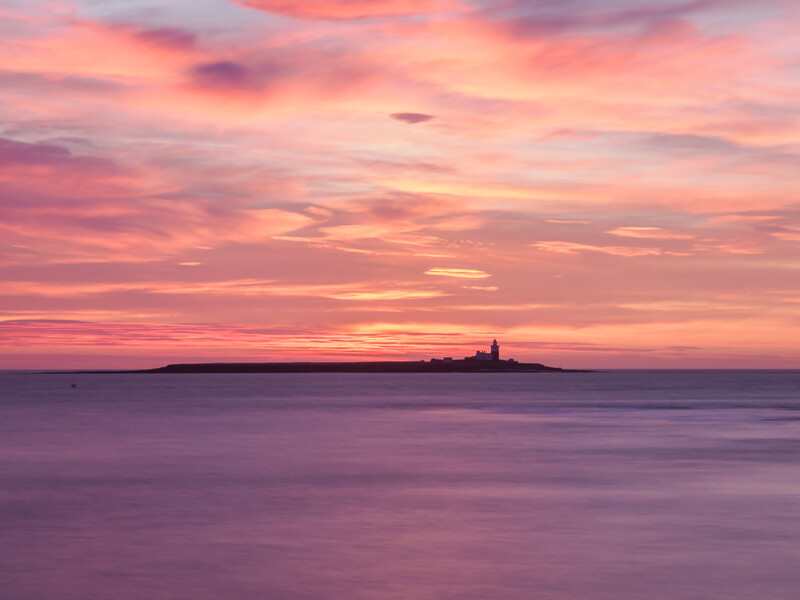 Silhouetted lighthouse on a distant island at sunset with pink and orange sky reflected in calm water.