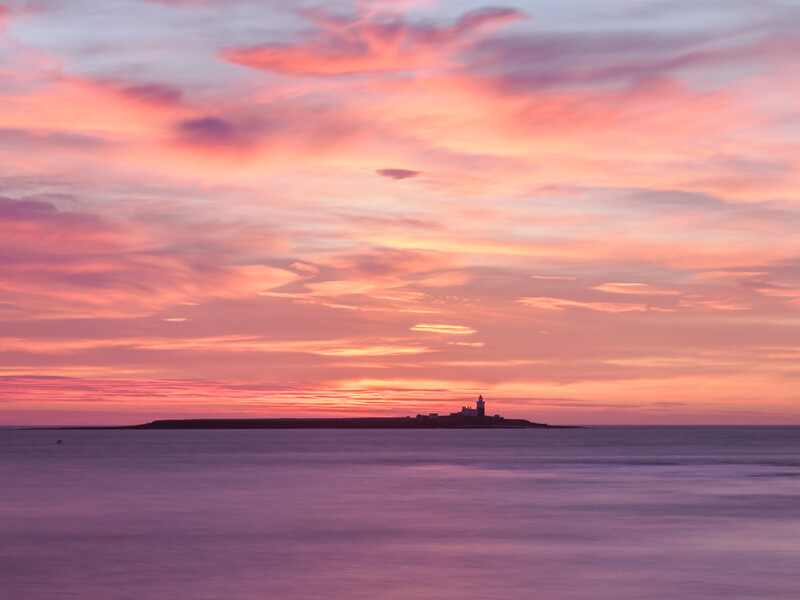 Silhouetted lighthouse on a distant island at sunset with dramatic pink and orange sky reflected in calm water.