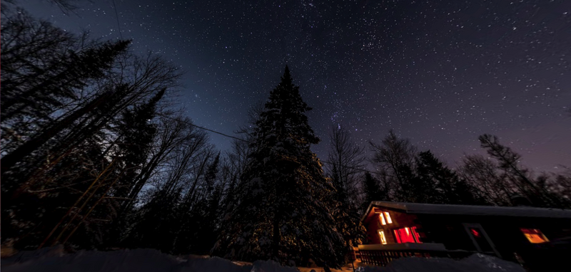 Snow-covered forest at night with warm-lit cabin and starry sky visible through the trees.