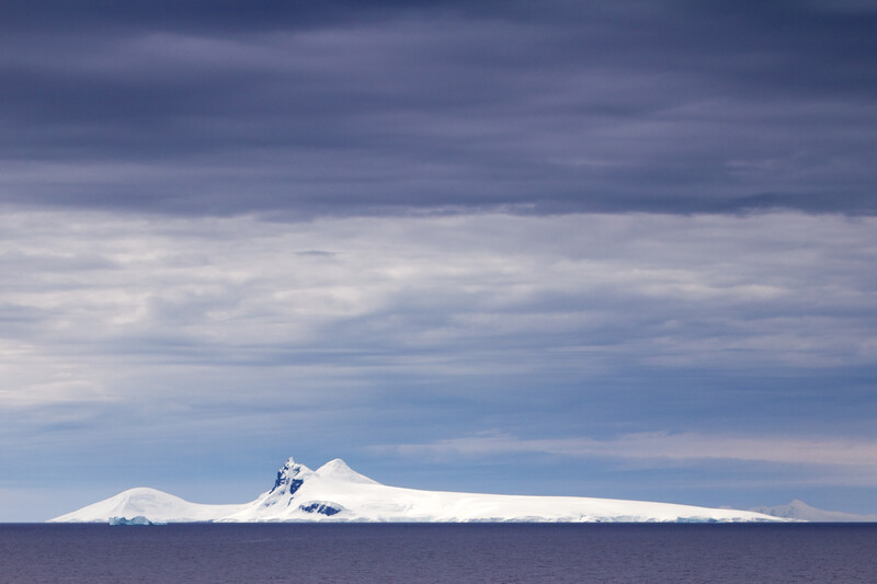 Snow-capped volcanic peak rising from calm ocean waters beneath a dramatic cloudy sky.