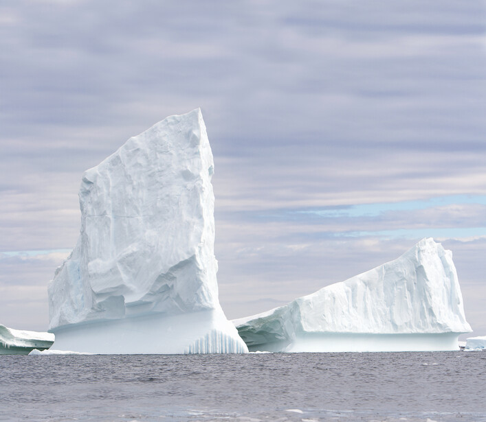 Two large icebergs with jagged peaks floating in calm ocean water under a cloudy sky.
