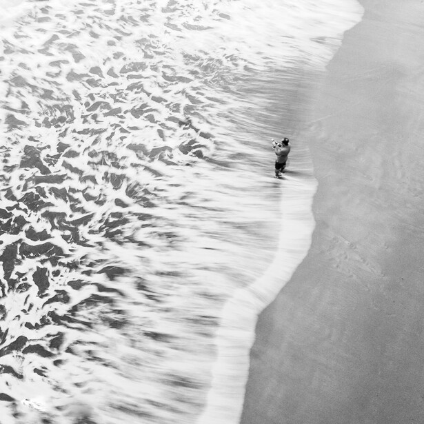 Lone surfer walking into ocean waves in black and white aerial perspective.
