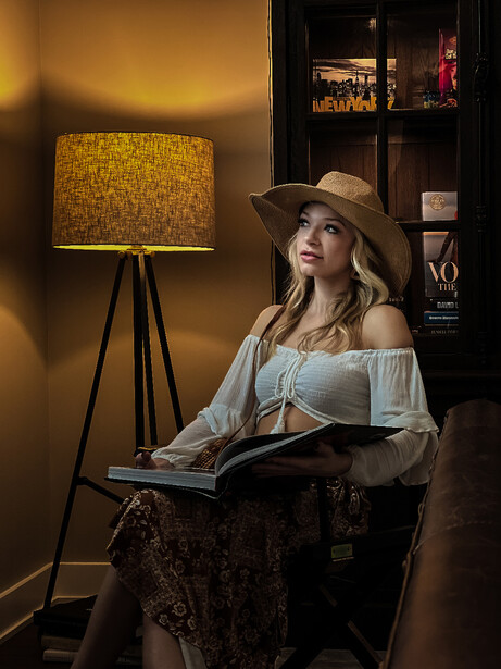Woman in off-shoulder blouse and wide-brimmed hat reading a book while seated in a chair.