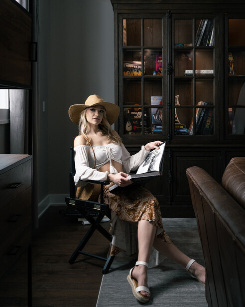 Woman in white shirt and straw hat reading magazine while seated in modern dark interior with bookshelves.