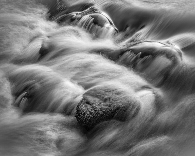 Long-exposure photograph of turbulent water flowing around a dark boulder, rendered in black and white.