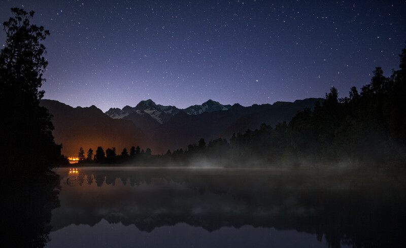 Mountain landscape at night with starry sky reflected in still lake water below silhouetted trees and a distant cabin light.